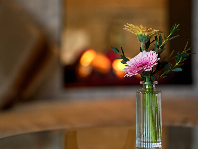 Tischdekoration im Hotel Oswald mit Blumen in einer Glasvase auf einem Glastisch, gemütlichem Sessel und knisterndem Kaminfeuer.