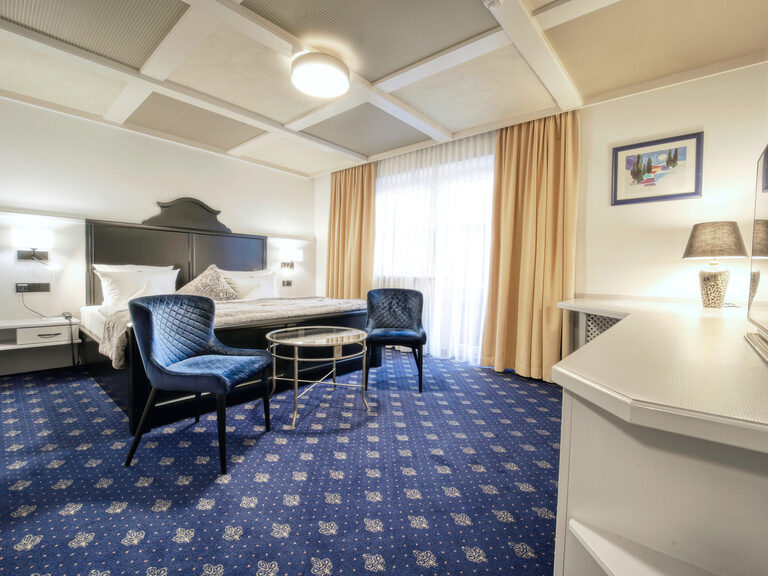 Das komfortable Doppelzimmer Roulette mit blauen Teppichboden, eleganter Sitzgelegenheit und viel Tageslicht im Hotel Oswald Bayerischer Wald.