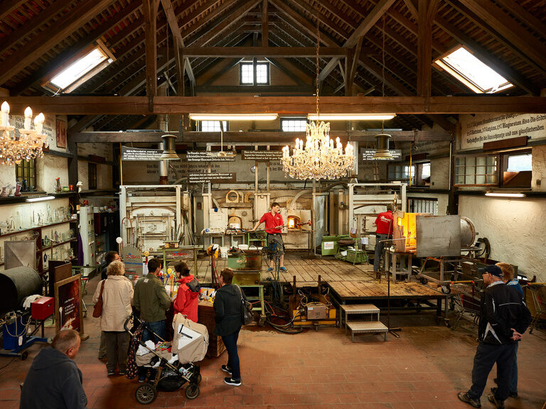 Besucher beobachten eine Glasvorführung in einer historischen Glashütte mit Kronleuchtern und Werkbühne im Bayerischen Wald.