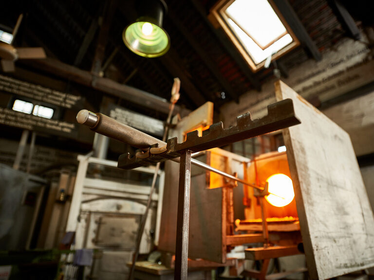 Nahaufnahme eines Glasofens mit leuchtendem Glasrohling auf einer Pfeife, Werkstattkulisse mit Licht und Werkzeugen im Hintergrund.