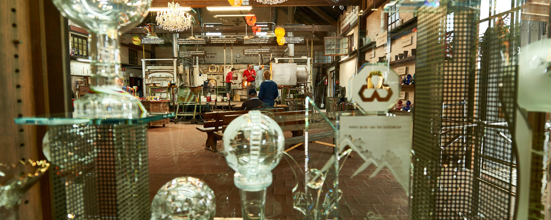 Blick durch ein Regal mit Glasobjekten in eine Werkstatt mit Glasöfen und Handwerkern bei der Arbeit, traditionelles Handwerk im Bayerischen Wald.