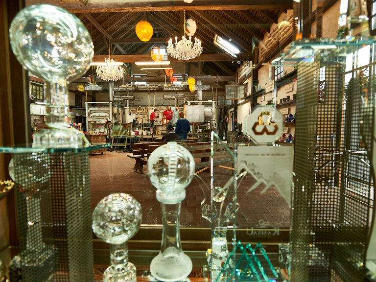 Blick durch ein Regal mit Glasobjekten in eine Werkstatt mit Glasöfen und Handwerkern bei der Arbeit, traditionelles Handwerk im Bayerischen Wald.