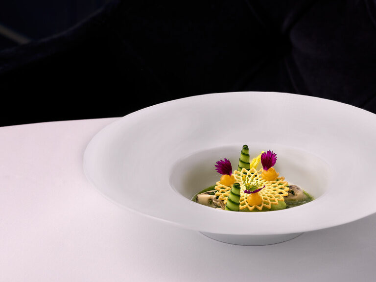 Kunstvoll angerichtetes Gourmetgericht auf einem tiefen, weißen Teller mit einer kunstvoll geschnittene gelbe Garnitur mit filigranem Muster im Hotel Oswald.