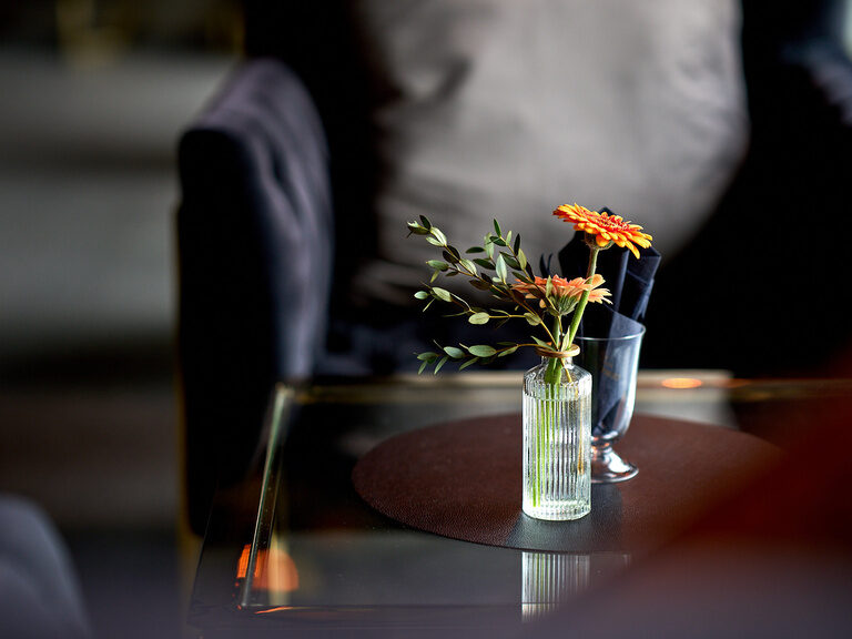 Tischdekoration im Hotel Oswald mit frischen Blumen in einer Glasvase auf einem edlen Glastisch, umgeben von gemütlichen Sesseln.