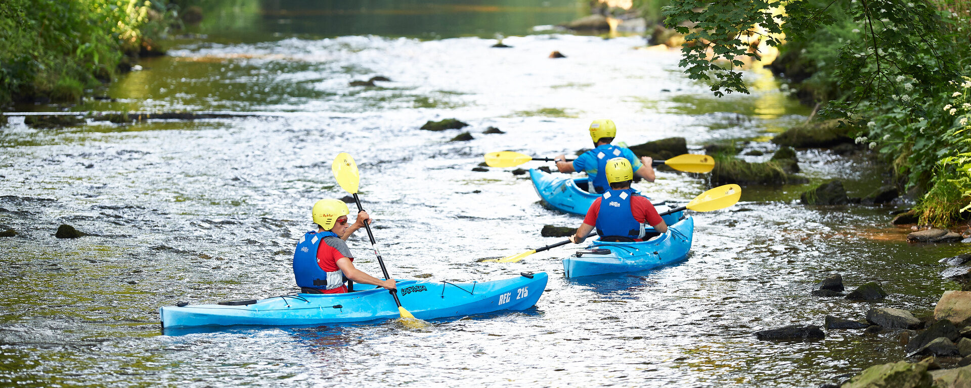 Gruppe von Kajakfahrern mit Schwimmwesten und Helmen auf einem ruhigen, bewaldeten Fluss im Bayerischen Wald.