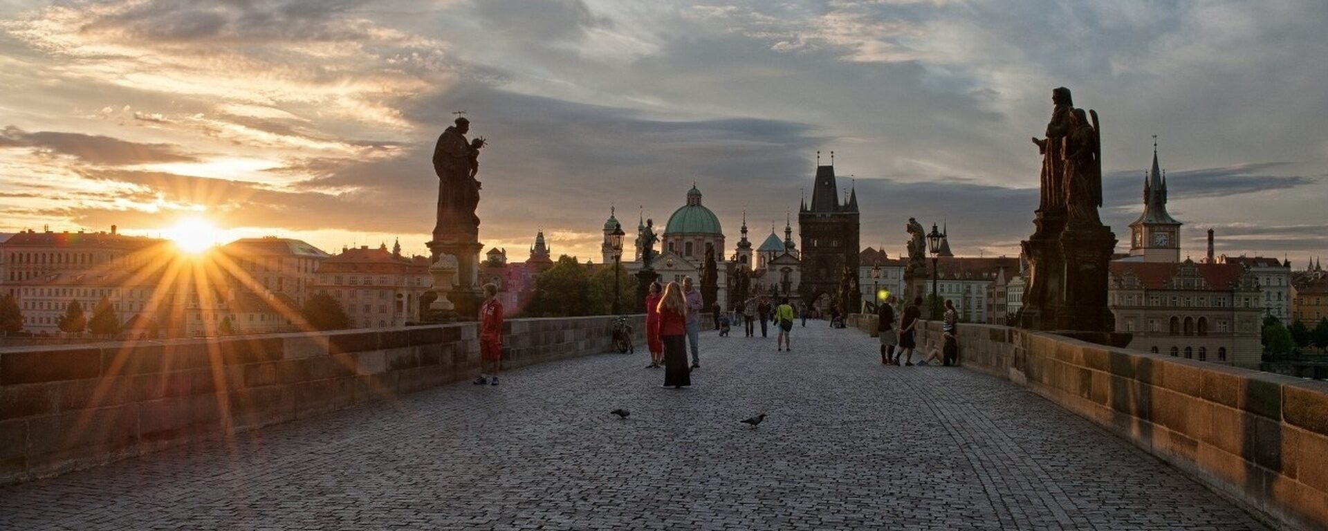 Karlsbrücke in Prag im Gegenlicht bei Sonnenaufgang mit wenigen Menschen und historischem Stadtpanorama im Hintergrund.