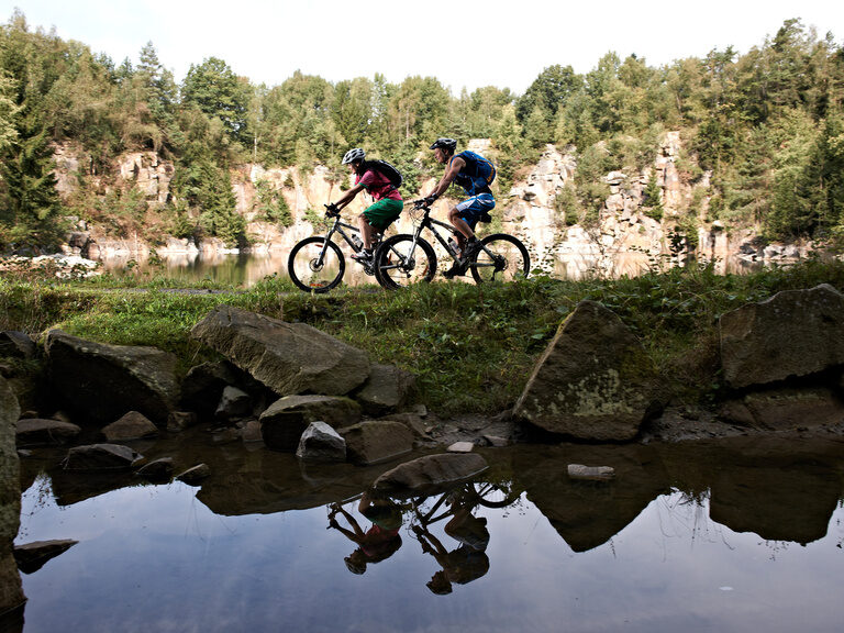 Zwei Radfahrer auf Mountainbikes fahren am Ufer eines Sees im Bayerischen Wald entlang, mit Felsen im Hintergrund.