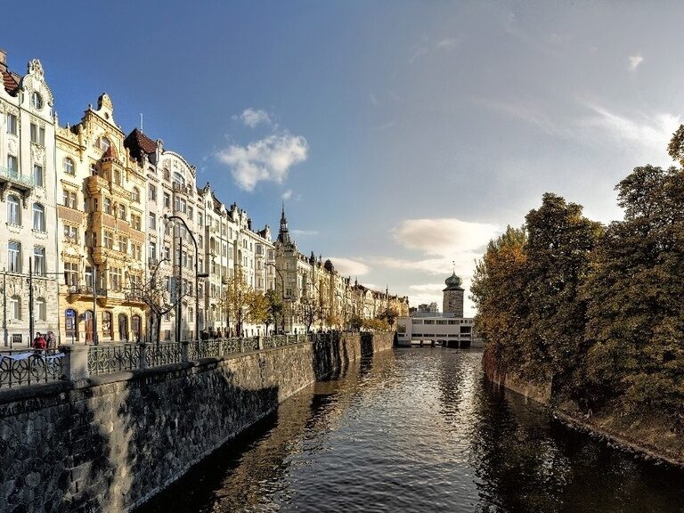 Uferstraße in Prag mit prächtigen Jugendstilgebäuden und Wasserlauf unter blauem Himmel.