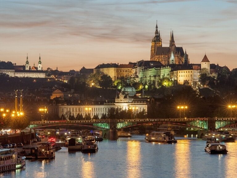 Abendliche Aussicht über die Moldau in Prag mit beleuchteter Prager Burg und Brückenverkehr auf dem Wasser.