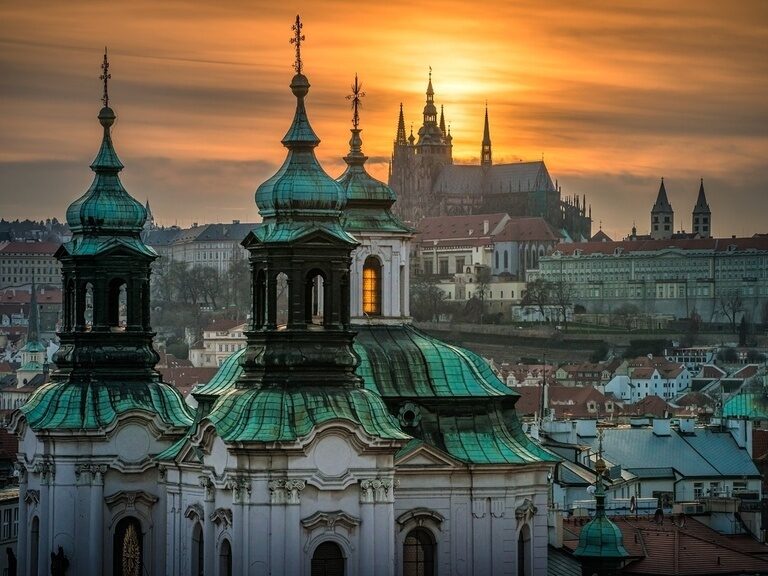 Sonnenuntergang hinter den Türmen einer Kirche in Prag, im Hintergrund die Prager Burg in warmem Licht.