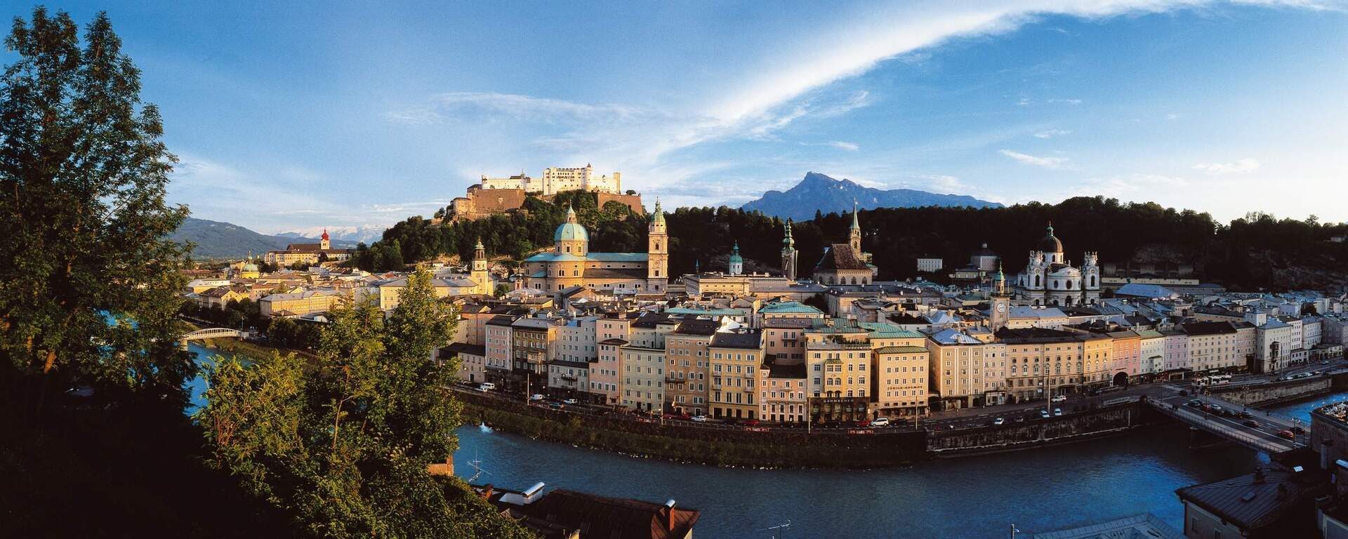 Panoramablick auf die Salzburger Altstadt mit der Festung Hohensalzburg und der Salzach. Dahinter ragt der Untersberg in den Himmel.