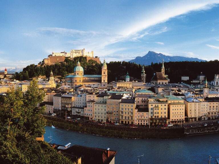 Panoramablick auf die Salzburger Altstadt mit der Festung Hohensalzburg und der Salzach. Dahinter ragt der Untersberg in den Himmel.