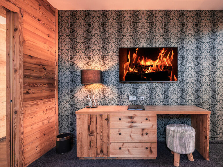 Die Suite  Jagasitz Plus im Hotel Oswald mit dunkel gemusterten Tapete und einem virtuellen Kamin für eine behagliche Atmosphäre.