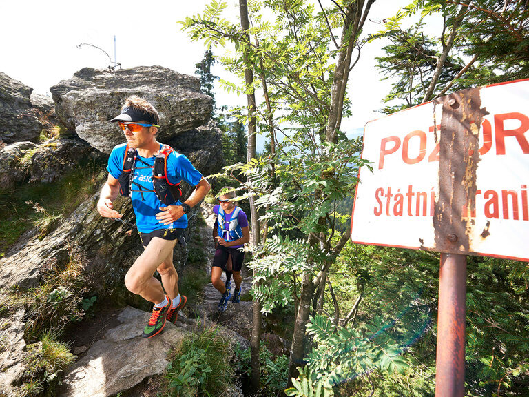 Zwei Trailrunner laufen über felsigen Grenzpfad bei einem Hinweisschild im Bayerischen Wald.