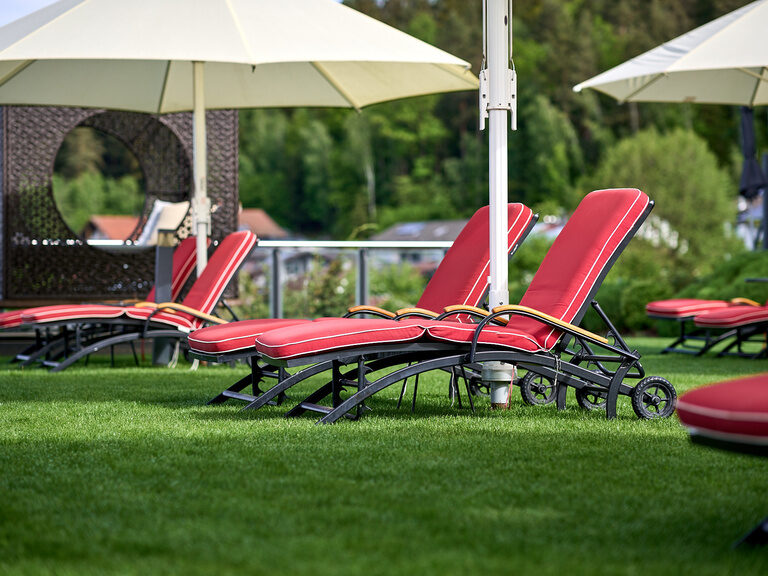 Der Wellnessgarten im Hotel Oswald mit bequemen roten Sonnenliegen und großen Sonnenschirmen in der gepflegten Gartenlandschaft