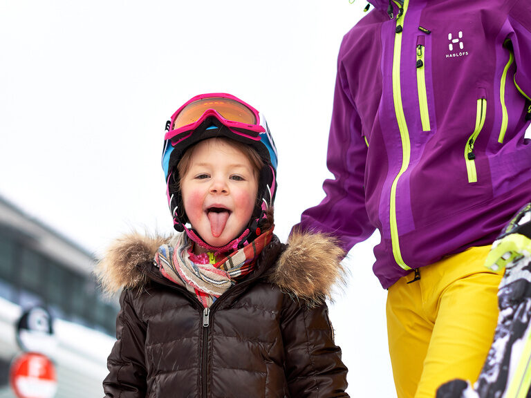 Ein Kind streckt lachend die Zunge heraus – bereit für einen Skitag mit seiner Mutter.
